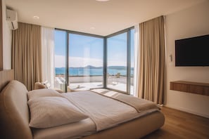 A modern hotel room with a large bed, a TV, and a balcony with a view of the sea.