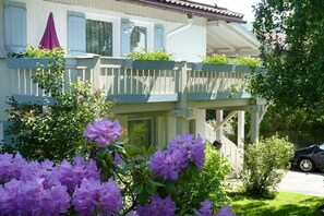 Ein zweistöckiges Gebäude mit Balkon, einem geparkten Auto davor und einem Garten mit violetten Blumen.