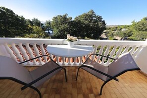 Ein Balkon mit rundem Tisch und zwei Stühlen, einem weißen Geländer und Blick auf Bäume und Gebäude.