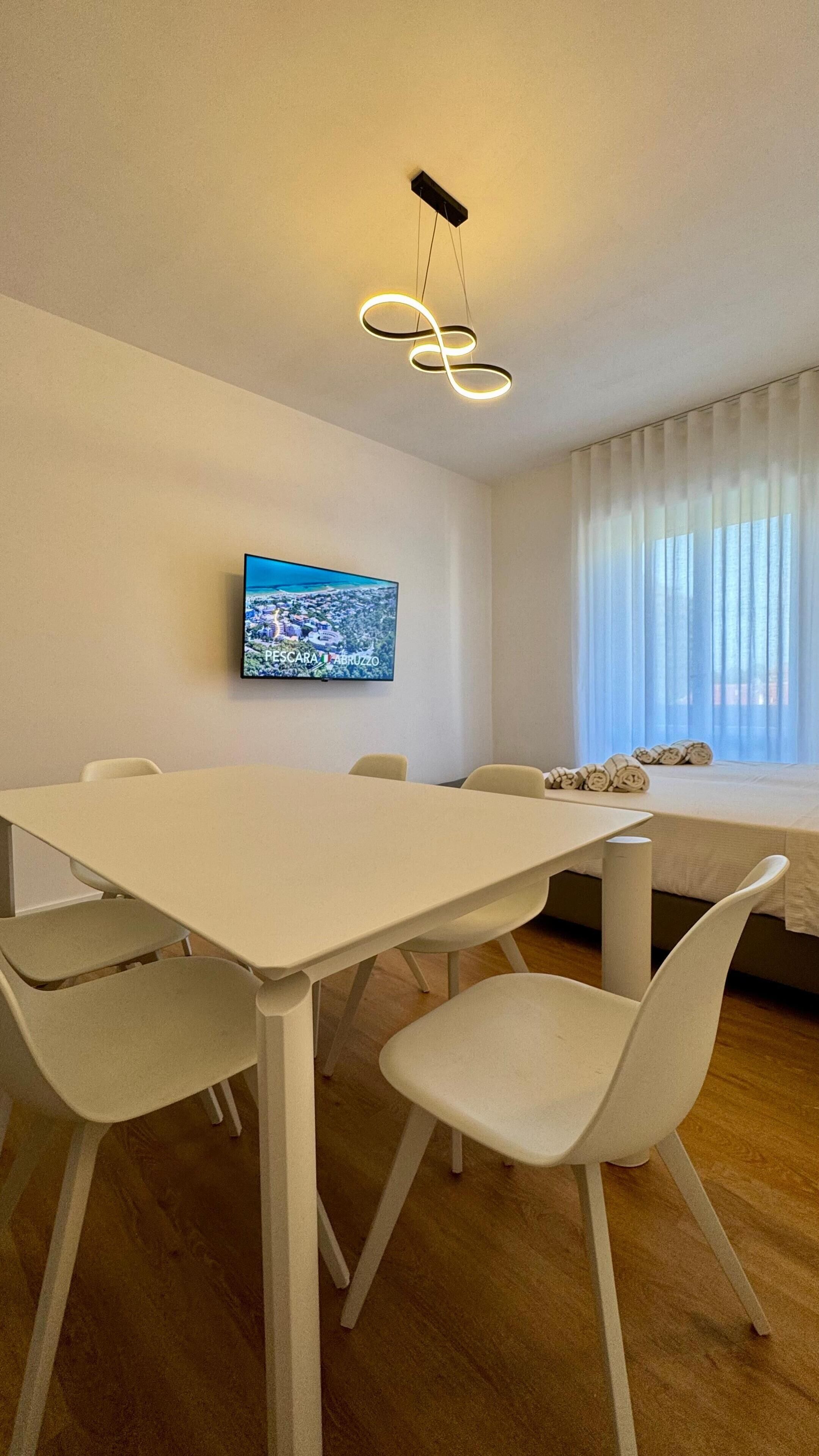 Ein moderner Essbereich mit einem weißen Tisch und Stühlen, einem Wandfernseher und einem Fenster mit durchscheinenden Vorhängen.