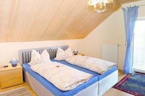 Ein Schlafzimmer mit schrägen Wänden, einem Bett mit floralem Bettbezug, einem Nachttisch mit Lampe und einem Fenster mit Vorhängen.