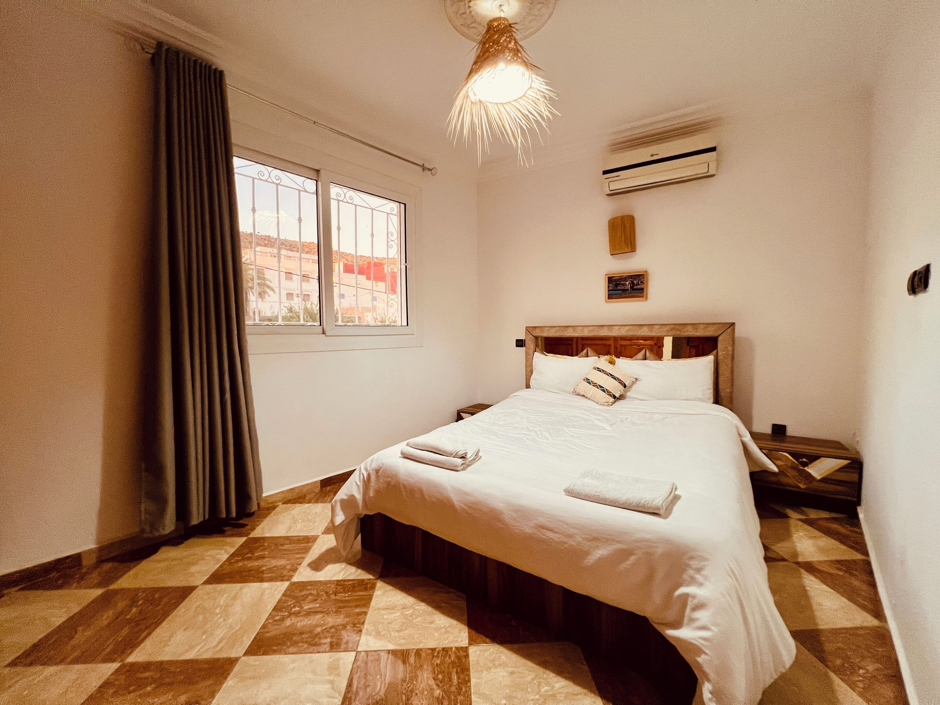 Un dormitorio con piso a cuadros, una cama con ropa blanca, un ventanal con cortinas, un equipo de aire acondicionado y una lámpara colgante.