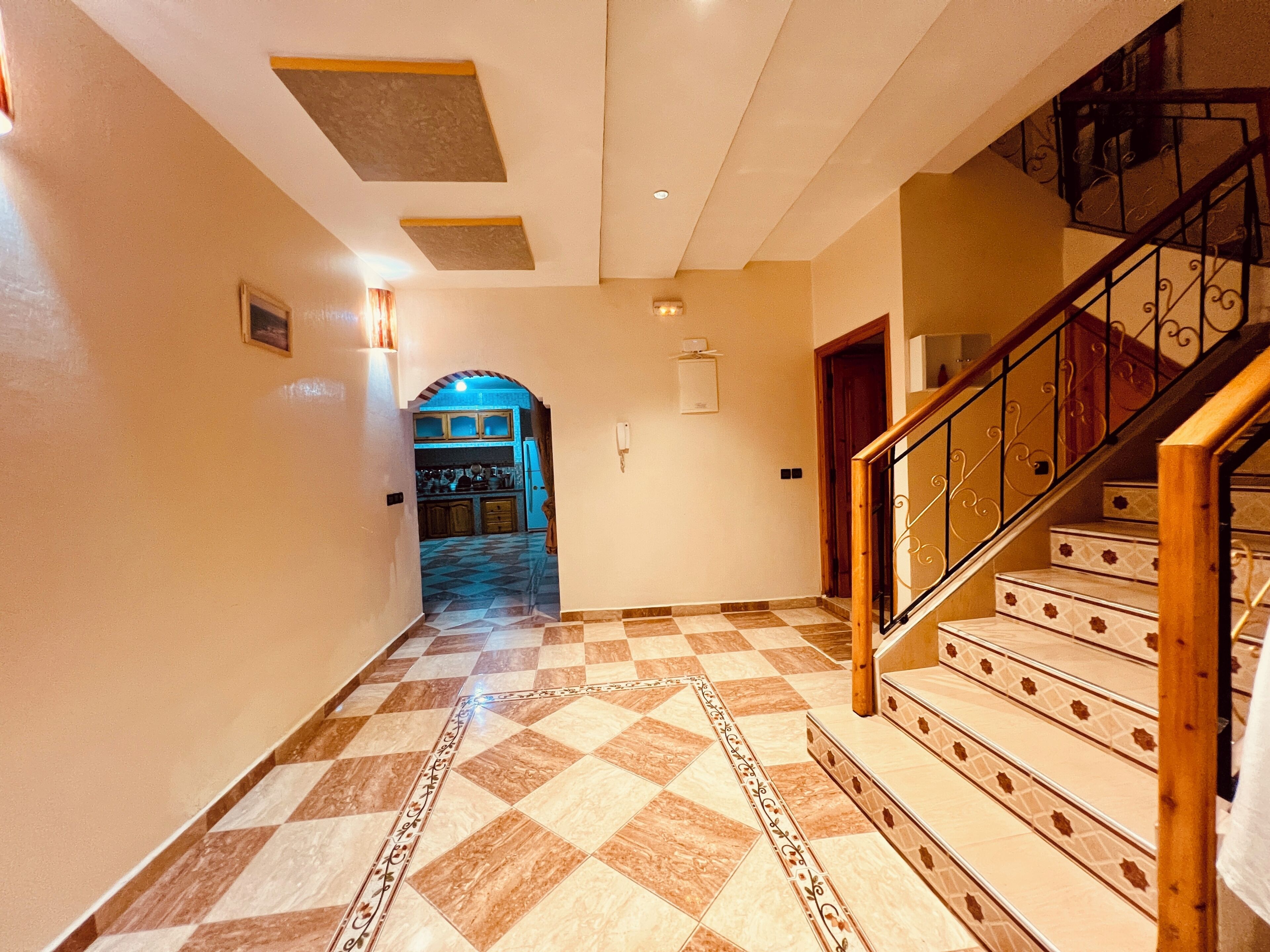 A hallway with a checkered floor, a staircase with wooden railings, and a kitchen visible through an archway.
