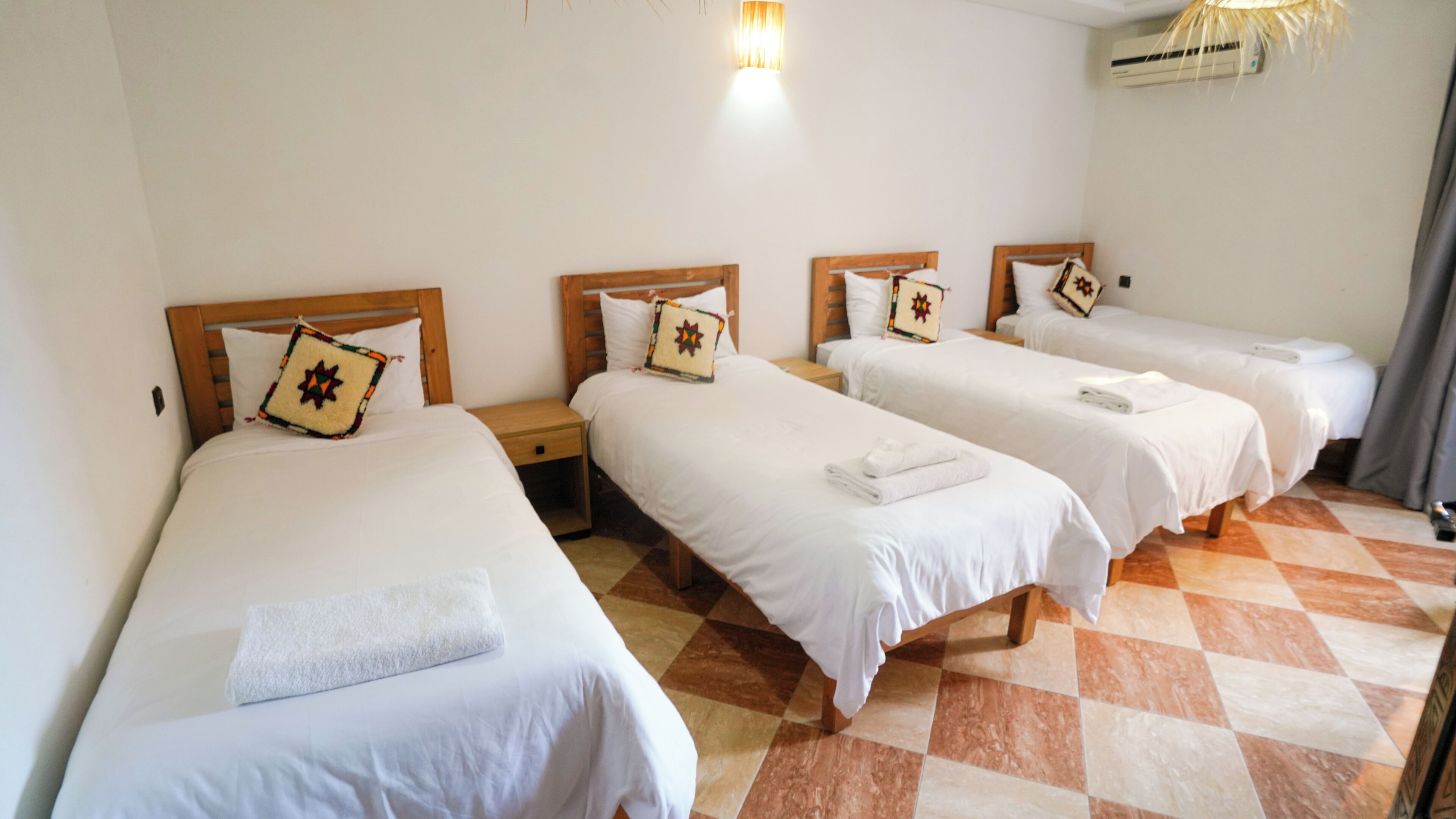 A hotel room with four single beds, each with a pillow and a white blanket. There is a nightstand between each bed, and a wall-mounted air conditioning unit above the beds.