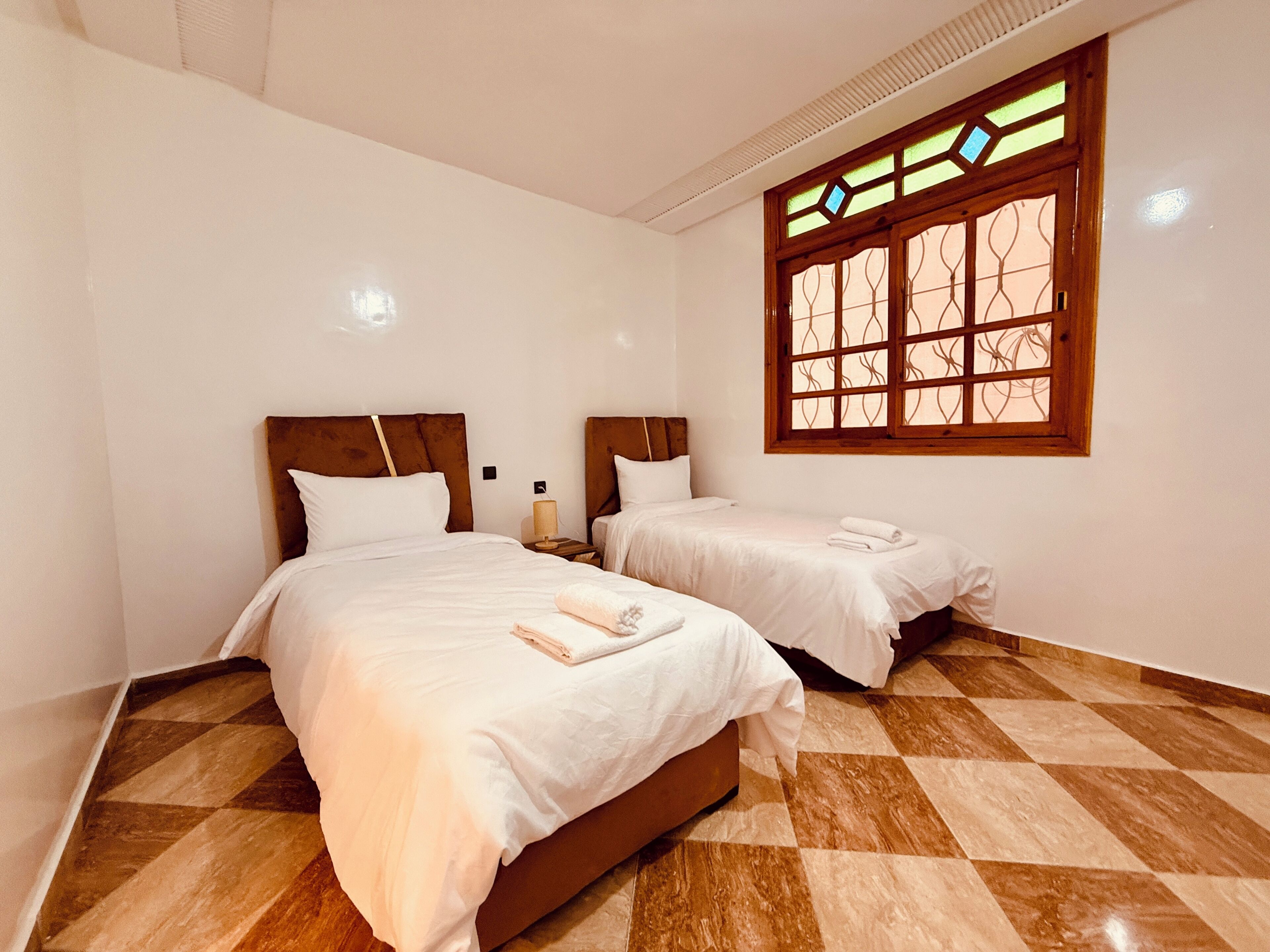 Quarto com duas camas, janela com padrão decorativo e chão quadriculado.