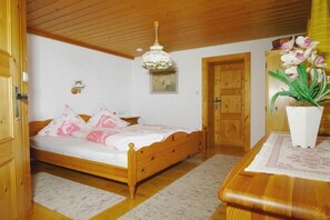 Ein hölzernes Bett mit weißer und pinkfarbener Bettdecke, ein Nachttisch aus Holz, eine Holzbalkendecke, eine Holztür, ein Holztisch mit weißem Tischlauf und ein Blumentopf mit weißen Blüten.