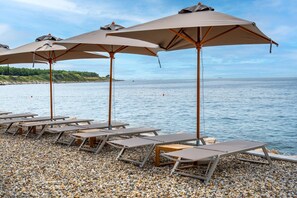 A pebbled beach with lounge chairs and umbrellas, overlooking calm water and a distant shoreline. - Deluxe Studio in Crveni Vrh (Crveni Vrh)