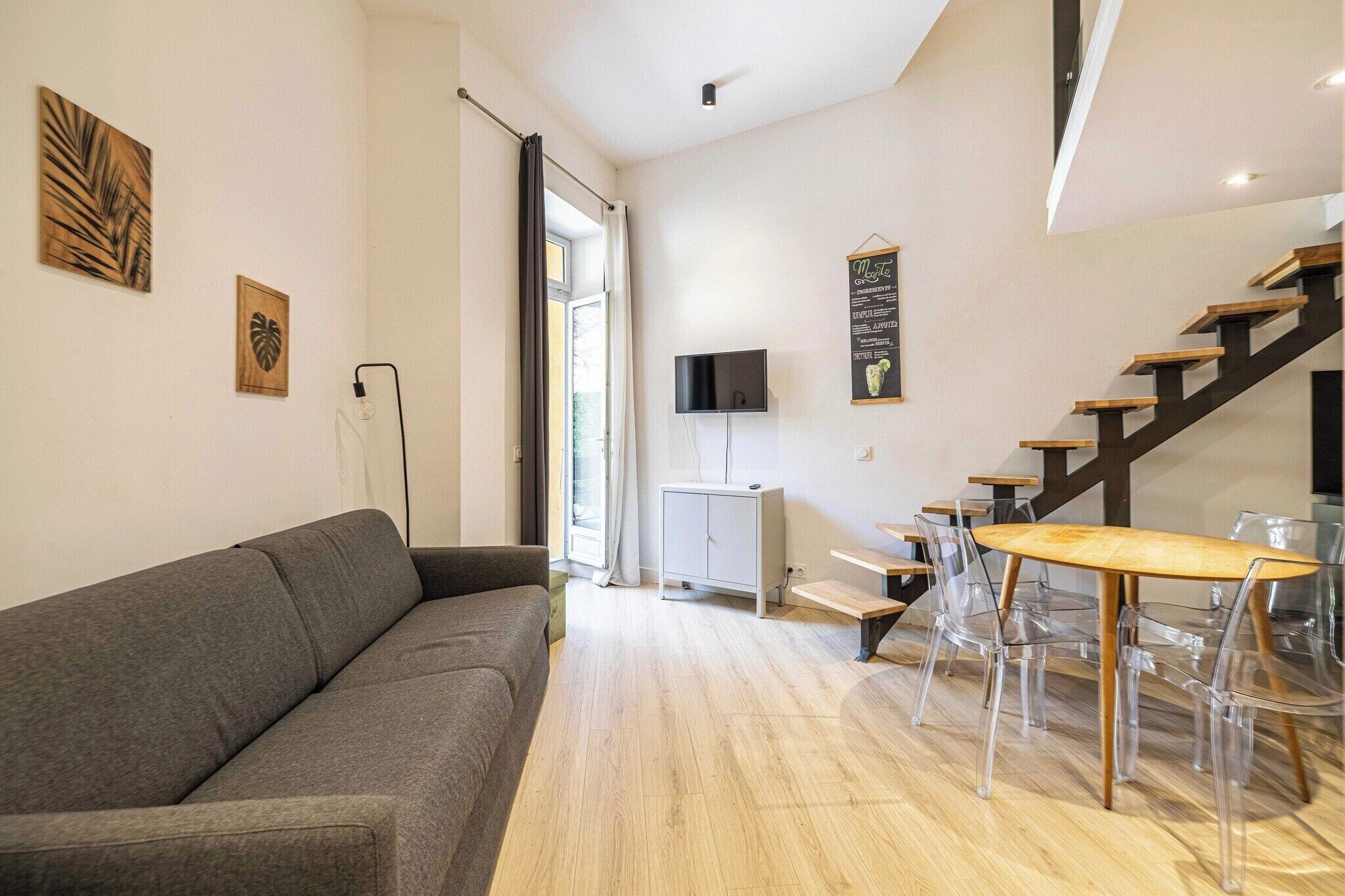 Ein modernes Wohnzimmer mit einem grauen Sofa, einem Esstisch mit transparenten Stühlen, einem Fernseher an der Wand und einer Treppe.