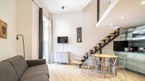 Een moderne woonkamer met een bank, een eettafel met stoelen, een trap, een keuken en een aan de muur bevestigde televisie.