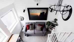 Uma sala de estar moderna com um sofá cinza, uma televisão montada, uma escada branca e uma área de jantar com mesa de madeira.