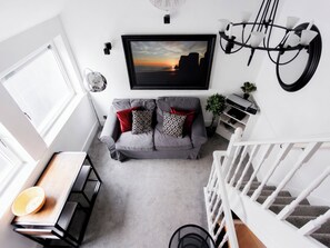 Ein modernes Wohnzimmer mit einem grauen Sofa, einem Wand-Fernseher, einer weißen Treppe und einem Essbereich mit einem Holztisch.