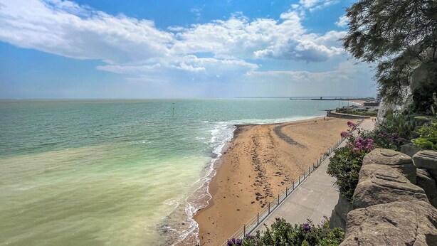 Uma praia costeira com céu claro, faixa de areia e um rochedo com vegetação.