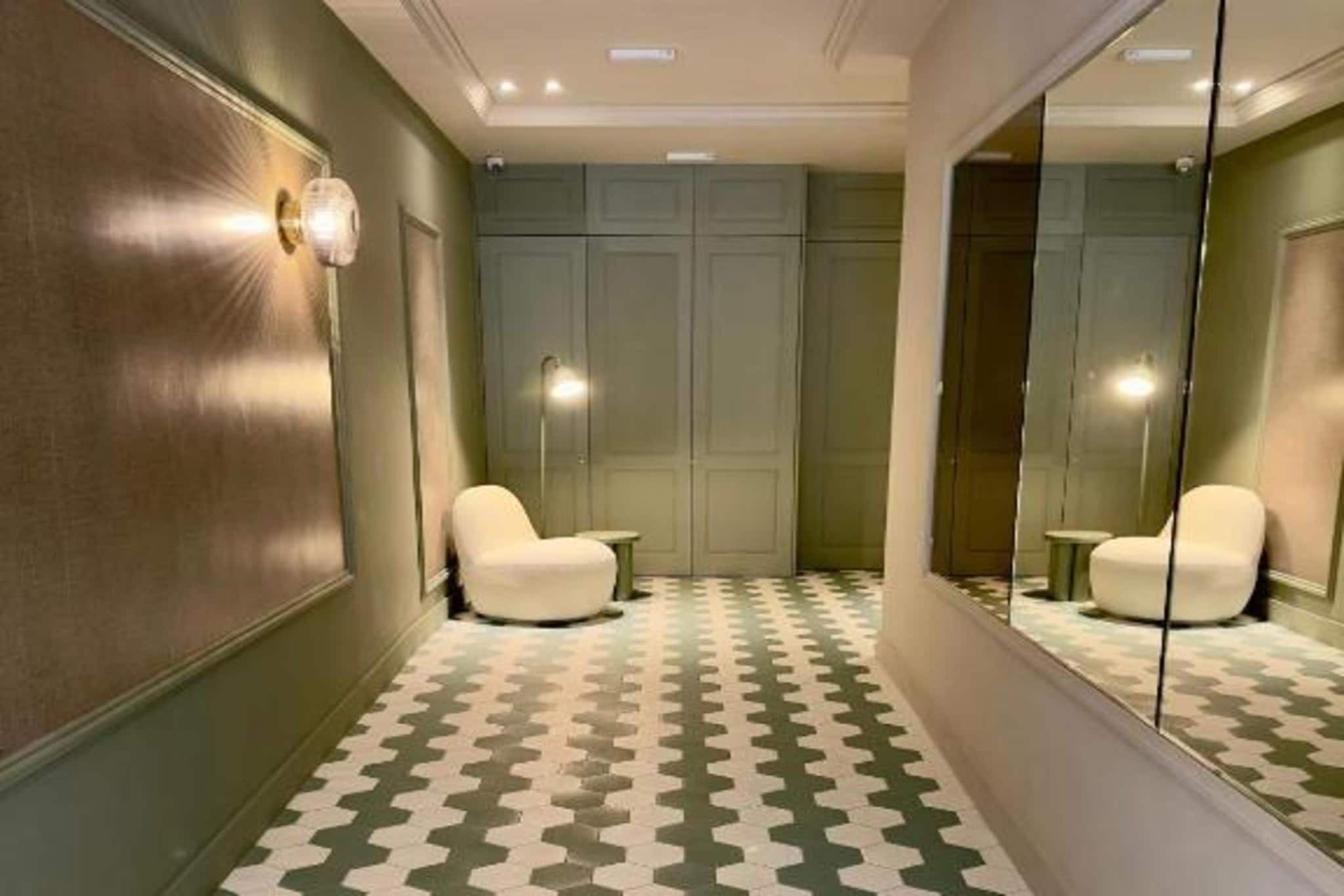 A hallway with a patterned floor, a mirror, and two white chairs.