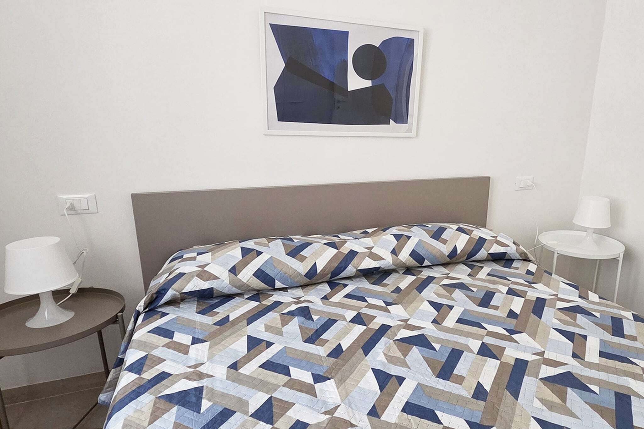 A hotel room with a bed, two bedside tables, a wall-mounted artwork, and a plain wall.