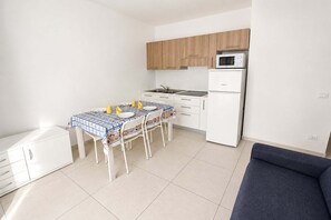 A compact kitchen with white cabinets, a microwave, a refrigerator, a dining table with chairs, and a sofa. - Appartment in Rosolina Mare (RO) (Rosolina Mare (RO))