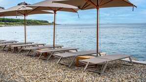 A pebbled beach with lounge chairs and umbrellas, overlooking calm water and a distant shoreline.