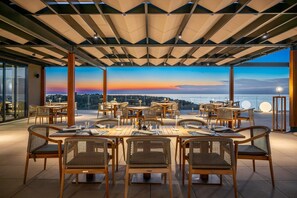 Un espacio al aire libre para comer con mesas y sillas, vista al horizonte de la ciudad y un atardecer.