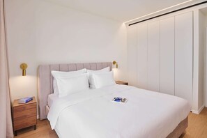 Un letto rifatto con lenzuola bianche, un comodino di legno con un libro e un lampadario a parete.
