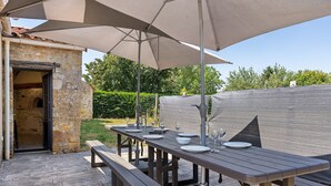 Un área de comedor al aire libre con un edificio de piedra, una mesa de madera dispuesta con platos y vasos, y un paraguas grande que da sombra.