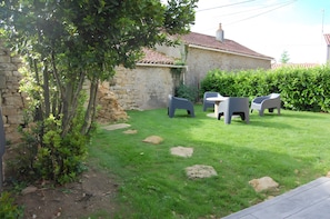 A stone building with a tiled roof, a grassy yard with a table and chairs, and a hedge.