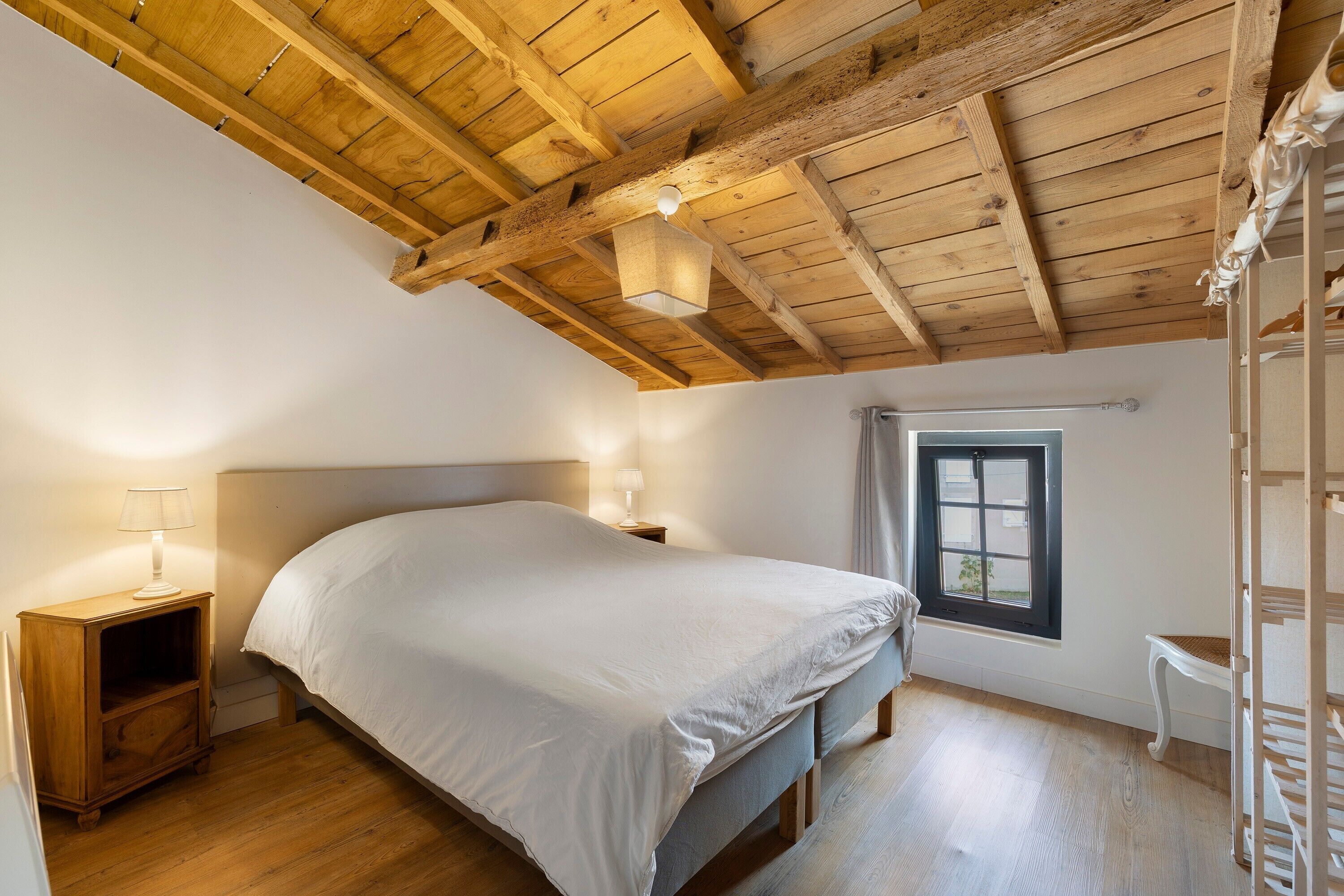 A bedroom with a wooden ceiling, a bed, a nightstand with a lamp, a window, and a chair.