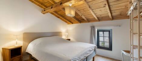 A bedroom with a wooden ceiling, a bed, a nightstand with a lamp, a window, and a chair.
