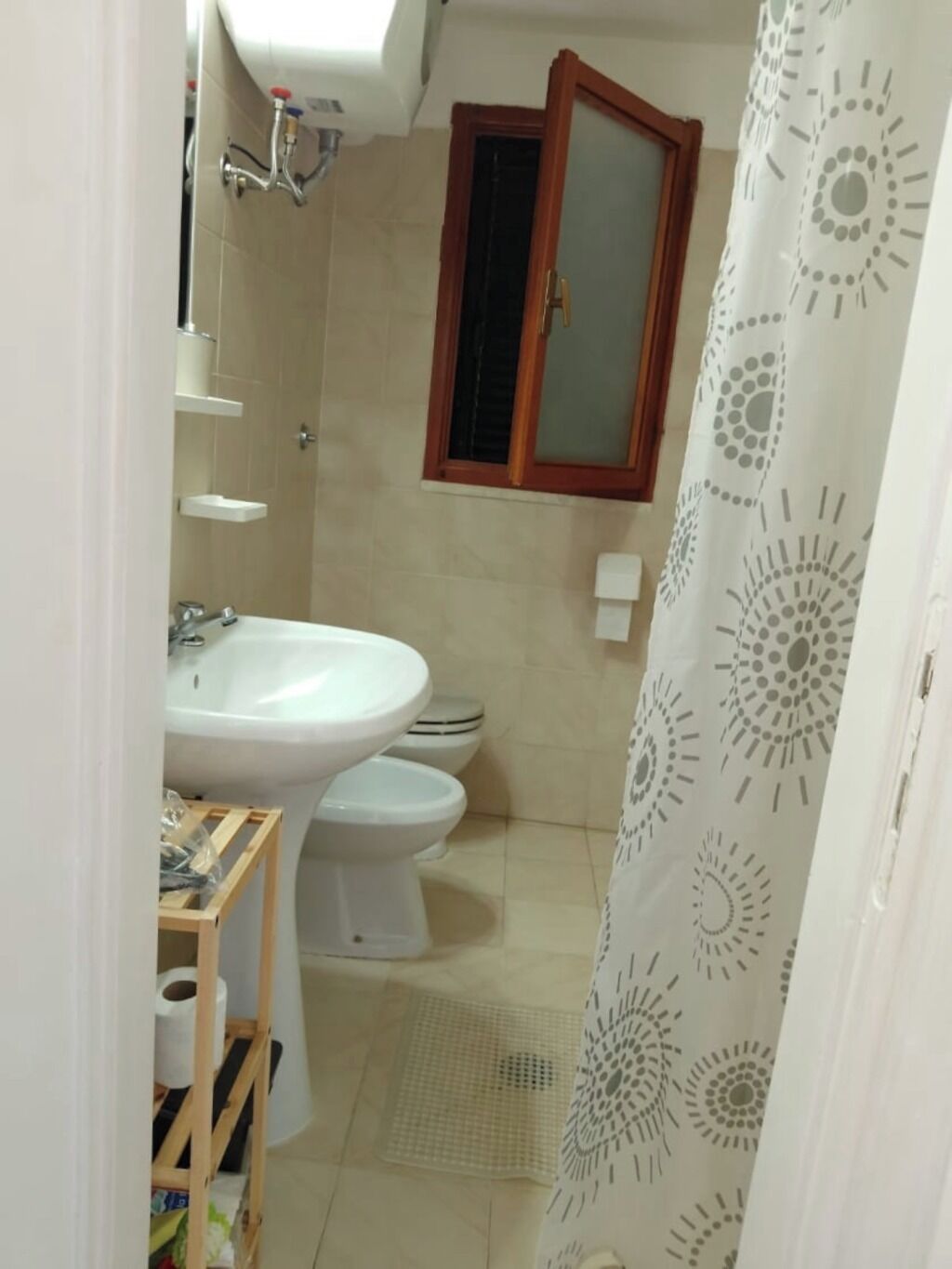 Un baño con lavamanos, dos inodoros, cortina de ducha y estante de madera con artículos de tocador.