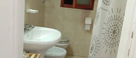 Un baño con lavamanos, dos inodoros, cortina de ducha y estante de madera con artículos de tocador.