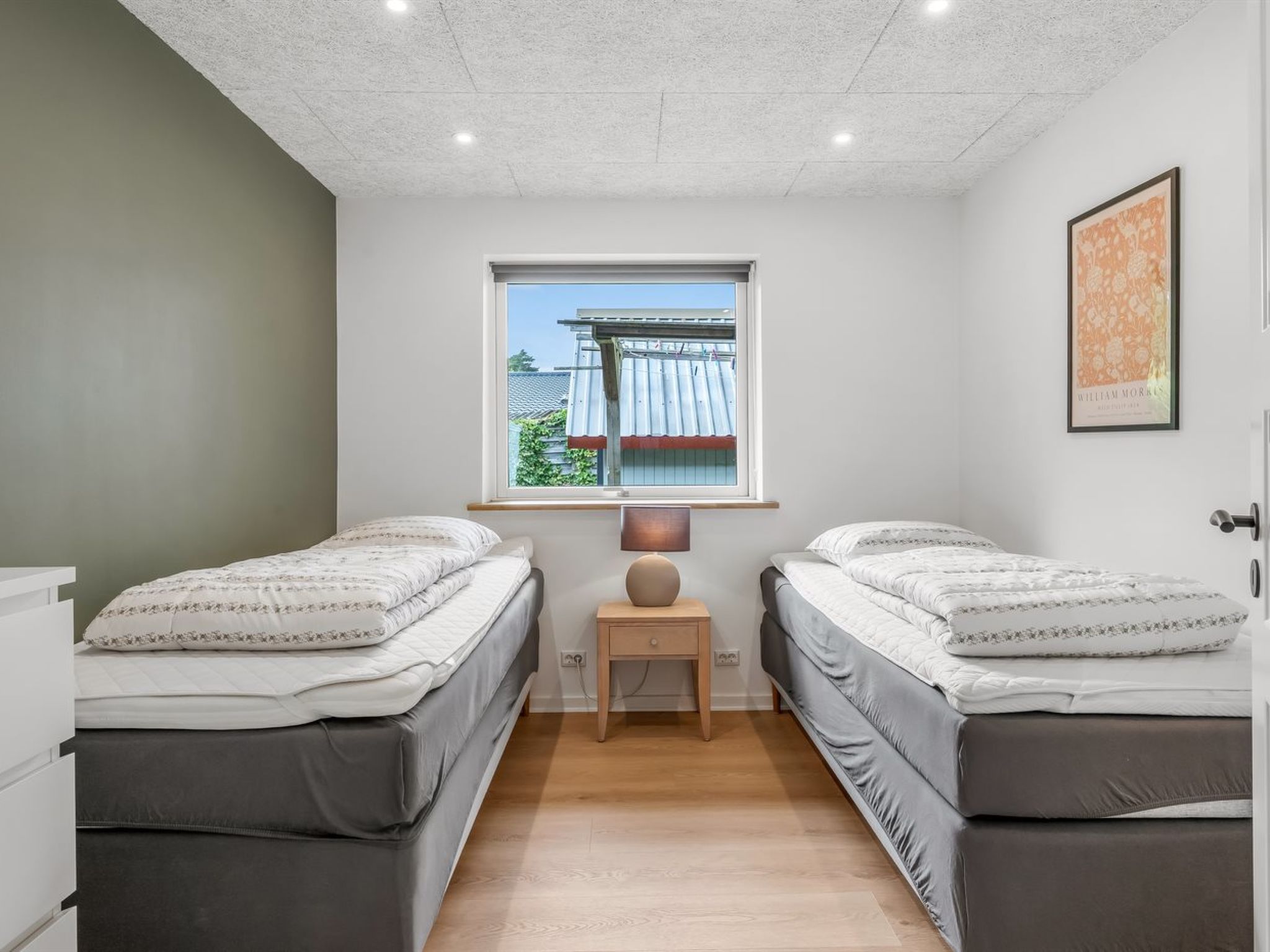 Une chambre avec deux lits, une fenêtre donnant sur une maison, une table de chevet avec une lampe et un tableau encadré au mur.