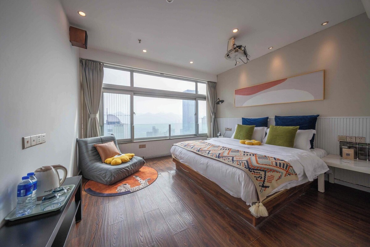 A modern hotel room with a large bed, a sofa, a desk with a laptop, and a view of the city.