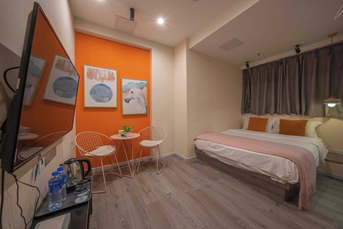 A hotel room with a bed, a desk with a chair, a small table with a plant, and artwork on the wall.