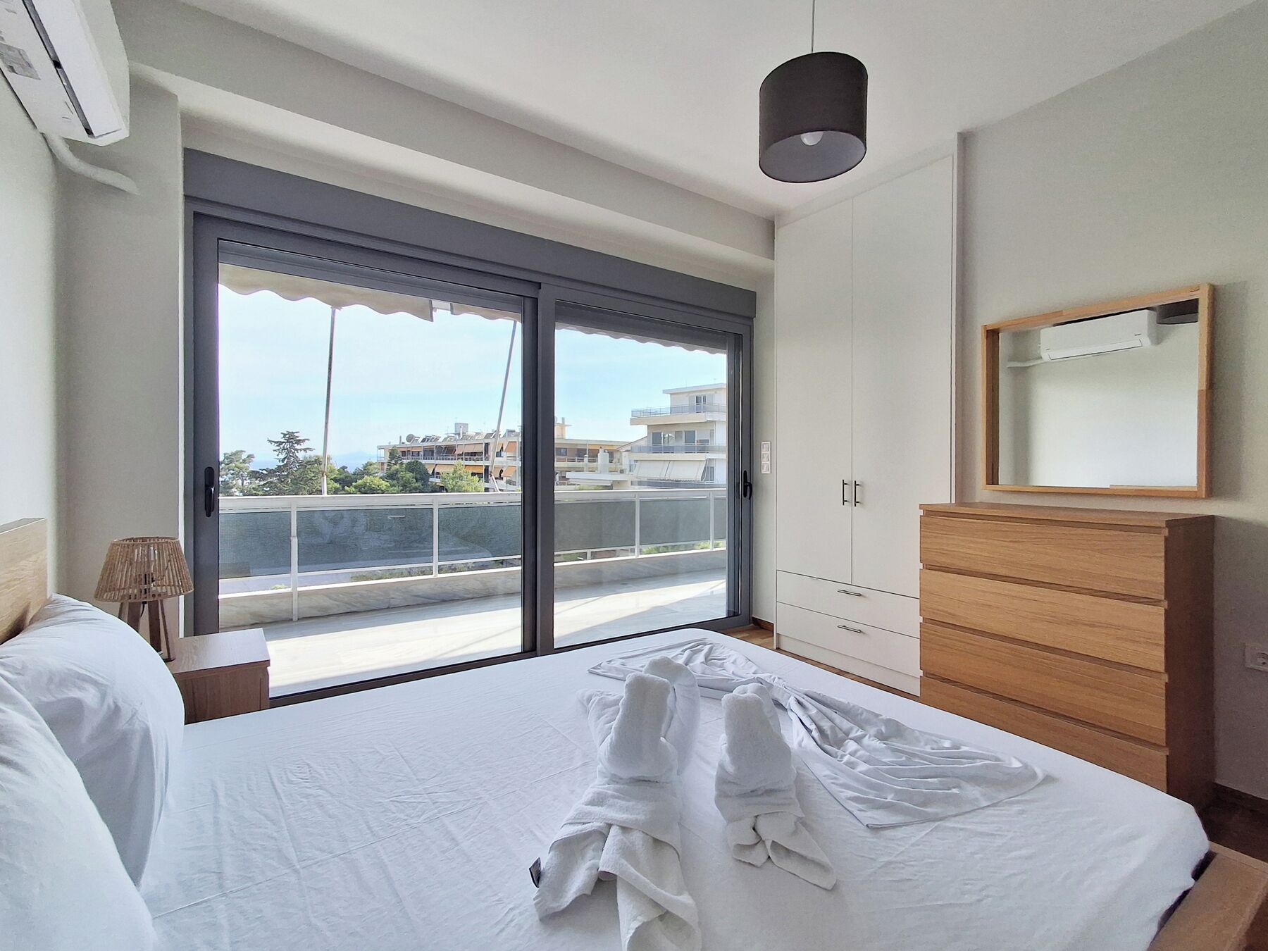 Un dormitorio con una cama, un aparador de madera, un espejo y un ventanal corredera a un balcón.