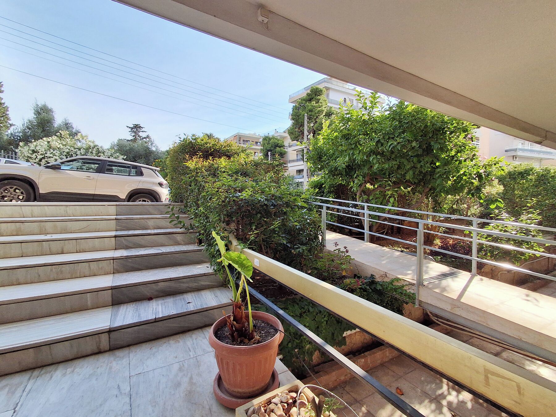 A balcony with a potted plant, a metal railing, and a view of a car and buildings in the background.