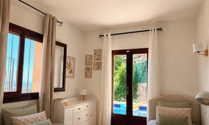 Ein Schlafzimmer mit Bett, Schminktisch, Nachttisch, einem Fenster mit Vorhängen, einer Tür zu einem Pool und Wandbildern.