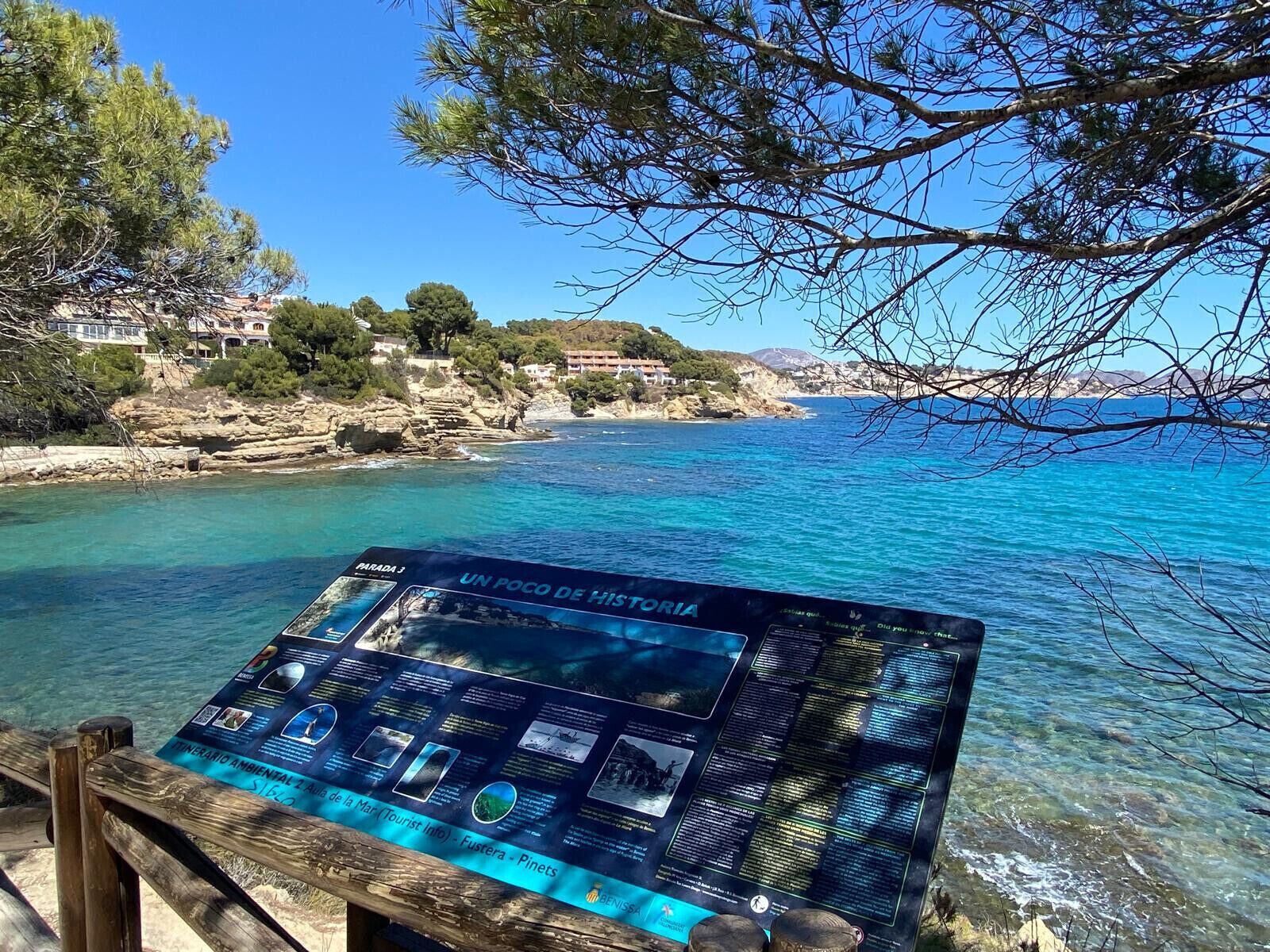 Une vue sur la côte, avec une mer bleue et limpide, un rivage rocheux et un panneau d’information en bois.