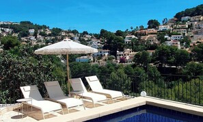 Ein Poolbereich mit Liegestühlen und Sonnenschirm, mit Blick auf einen Hügel mit Gebäuden.