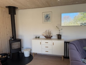 Ein gemütlicher Raum mit Kaminofen, einem weißen Schrank mit Schubladen, einem Schiffsmodell und einem Fenster mit Blick auf Bäume.