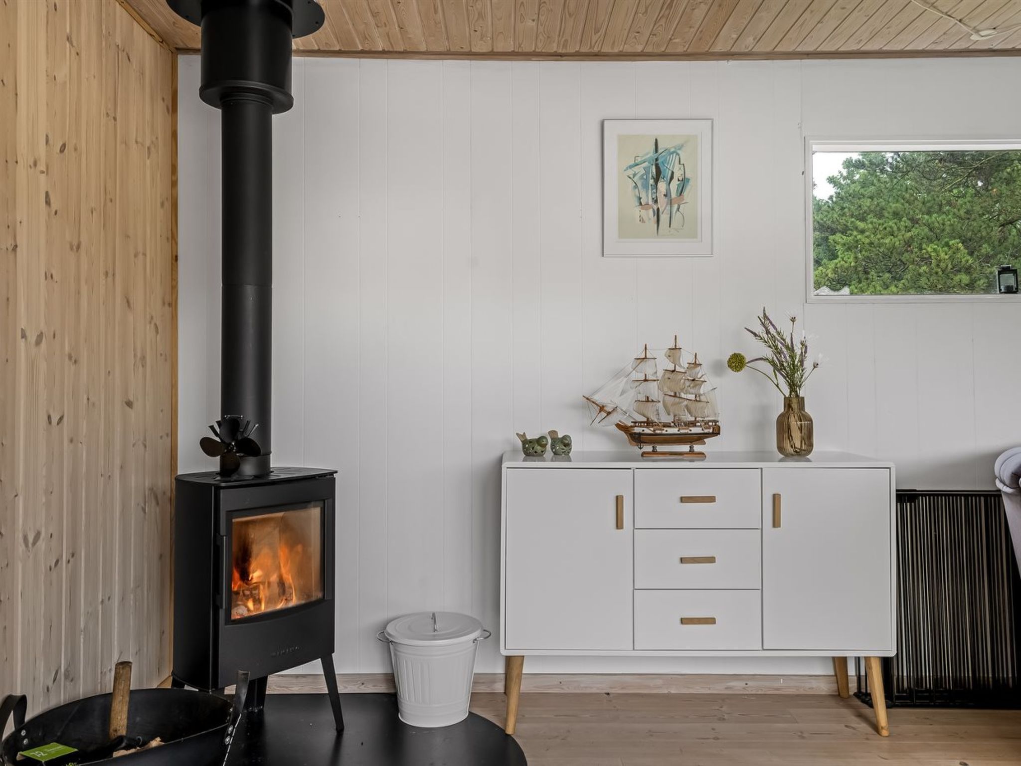 A room with a black wood-burning stove, white cabinetry, a small ship model, and a framed abstract painting.