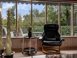 A room with a black leather chair, a small table with a vase of flowers, and a view of greenery through large windows. - "Haley" - 380m from the sea by Interhome (Hadsund)