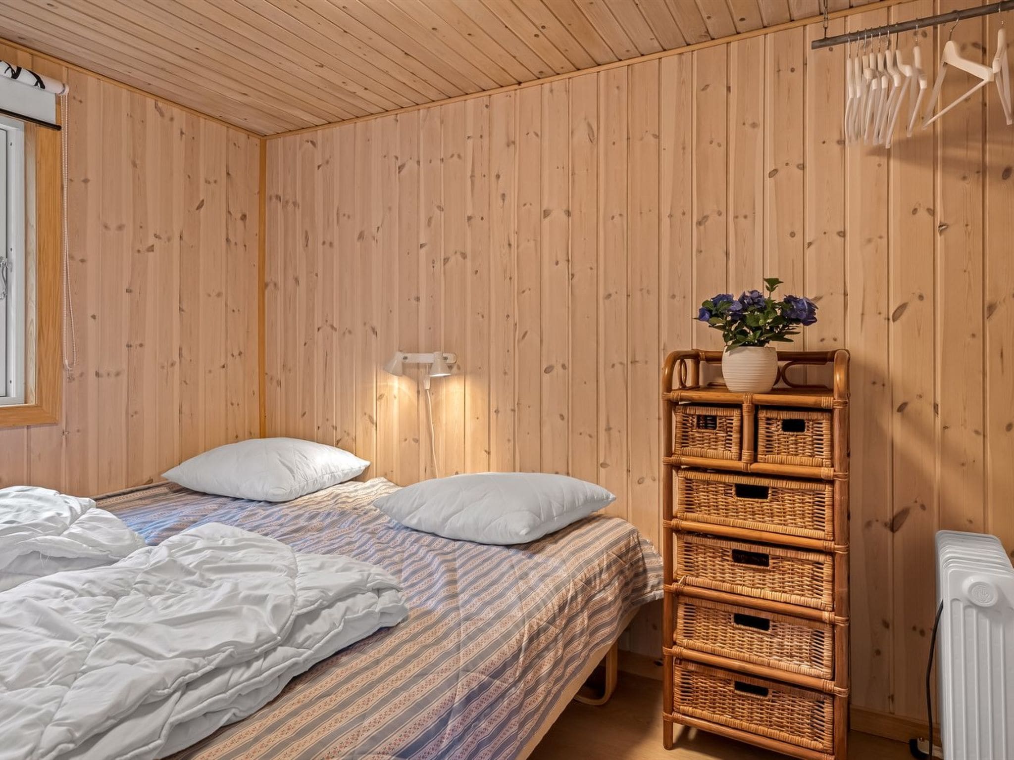 A bedroom with a bed, pillows, a wall-mounted lamp, a wicker storage unit, and a radiator.