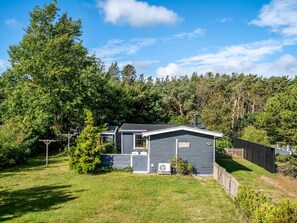 Une maison de plain-pied avec un petit garage attenant, entourée d’une cour clôturée et d’arbres touffus.