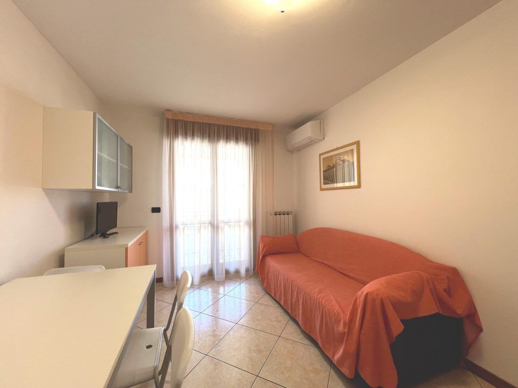 Une petite pièce bien éclairée, équipée d’un lit, d’un bureau, d’une chaise, d’un téléviseur et d’un radiateur.