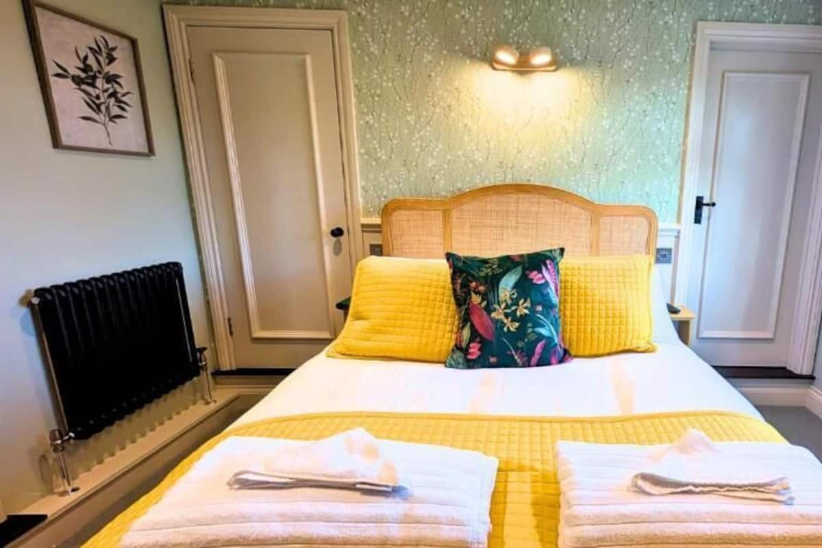 A bedroom with a bed, two pillows, a bedside table, a wall-mounted light, a radiator, and a window with curtains.