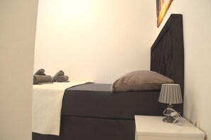 Ein Bett mit dunklem Kopfteil, einem Kissen, einer Nachttischlampe und zusammengerollten Handtüchern auf einem weißen Tisch.