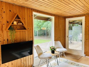 Ein Zimmer mit Holzwänden, einem Fernseher, einem kleinen Regal mit Spielzeug und Blick auf einen Garten durch eine Schiebetür.