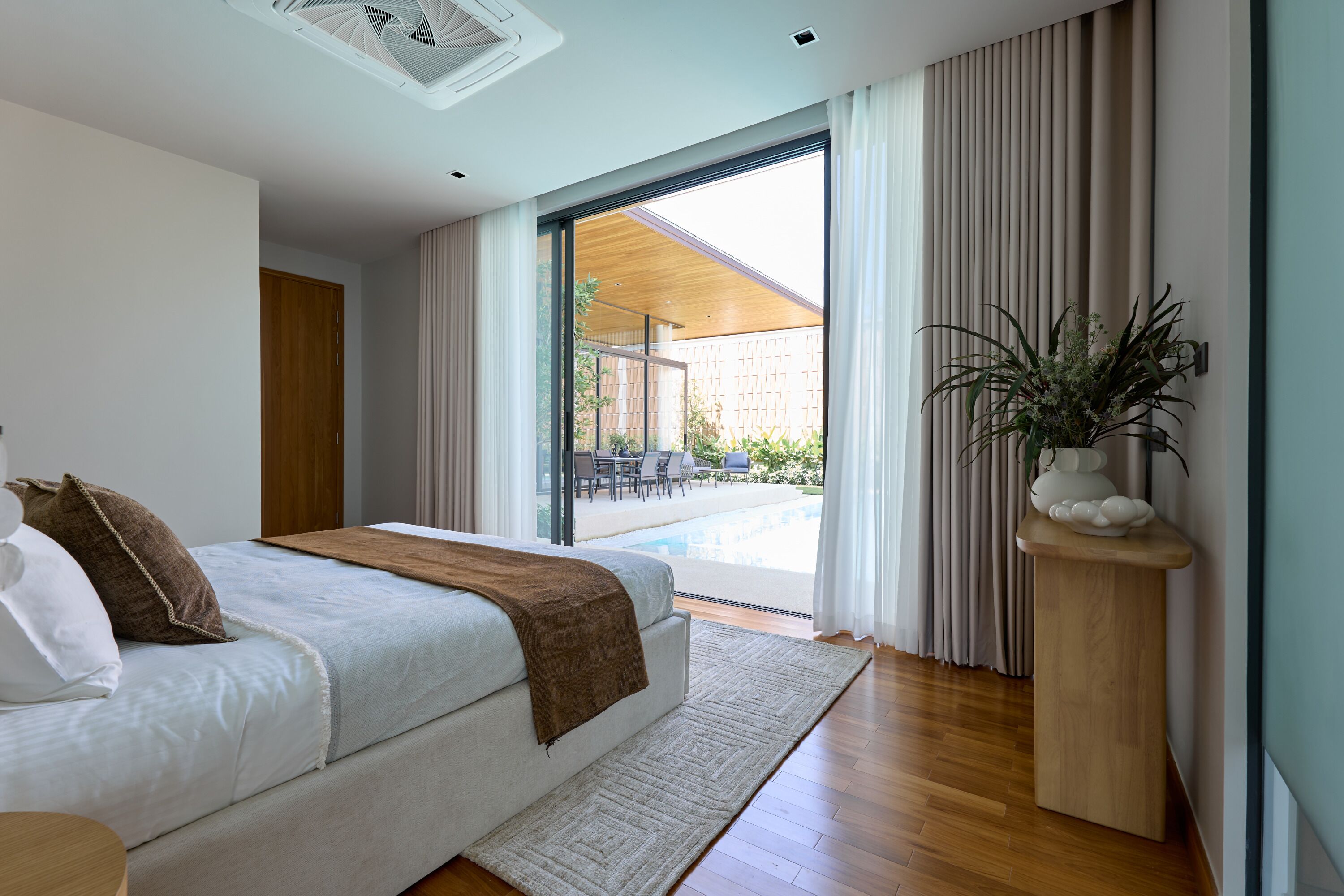 A modern bedroom with a large bed, a view of an outdoor pool, and a potted plant.