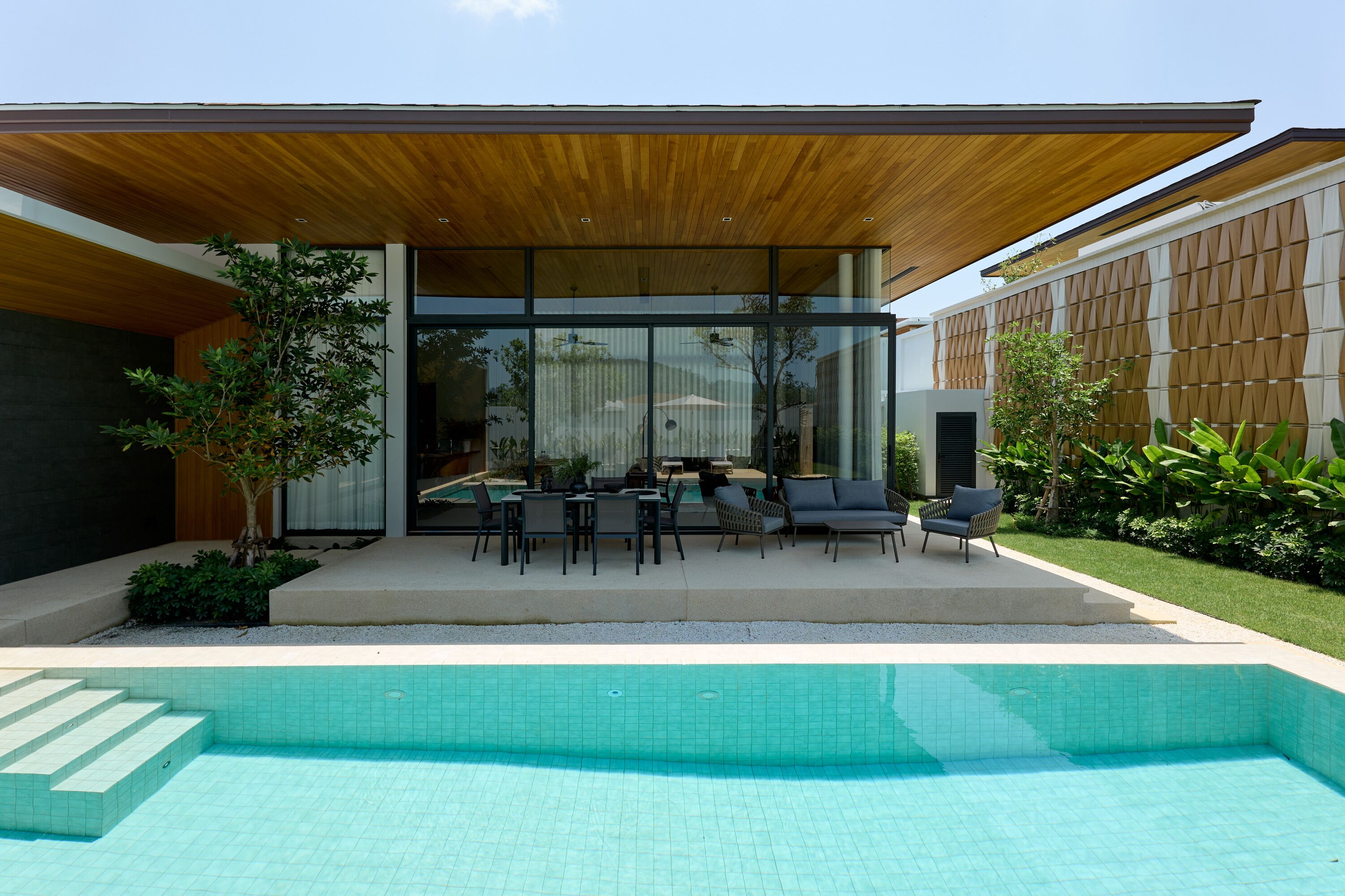 A modern outdoor pool area with a covered seating area, a pool, and surrounding greenery.