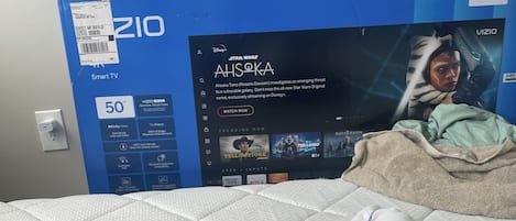 En säng med en vit, vadderad madrass, en person som ligger ner och en tv-box märkt med "VIZIO" samt en affisch med Star Wars-karaktären Ahsoka.