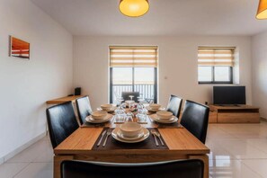 A dining area with a wooden table set for six, flanked by black leather chairs, with two windows and a television in the background.