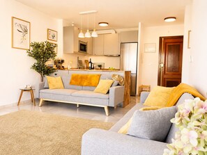 Ein modernes Wohnzimmer mit einem grauen Sofa, gelben Kissen, einem Holztisch und einer Küche im Hintergrund.